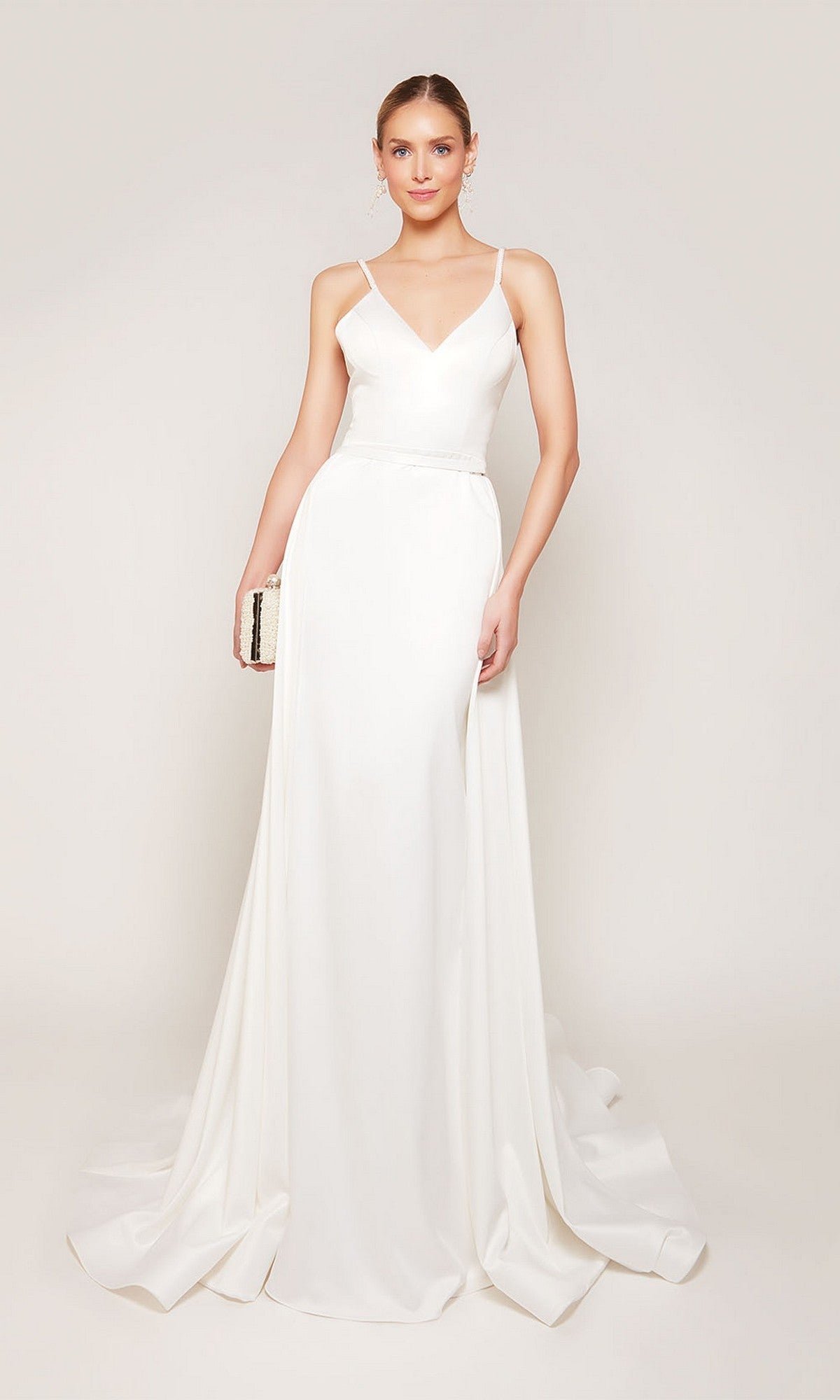 A woman in the Alyce 7090 long formal dress with thin straps and a detachable floor-length overskirt holds a small clutch, standing against a plain light background.